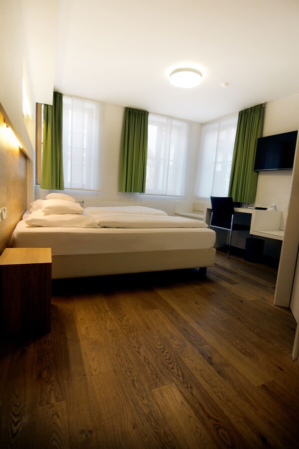 Una habitación de hotel moderna con suelo de madera, cama con ropa blanca, escritorio con silla y televisor de pantalla plana montado en la pared.