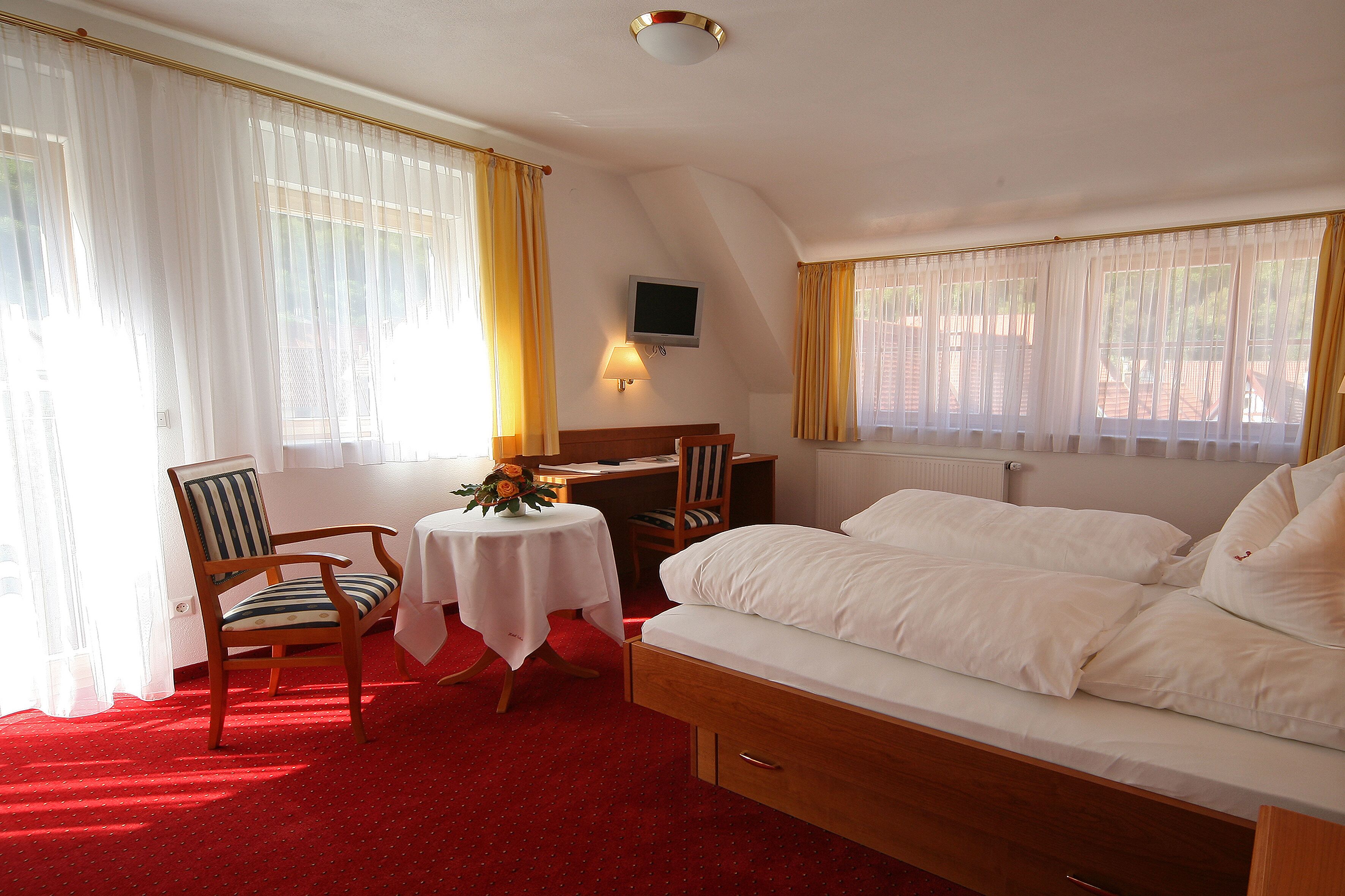 A hotel room with a bed, a desk, a chair, a table with a flower, and a TV on the wall.