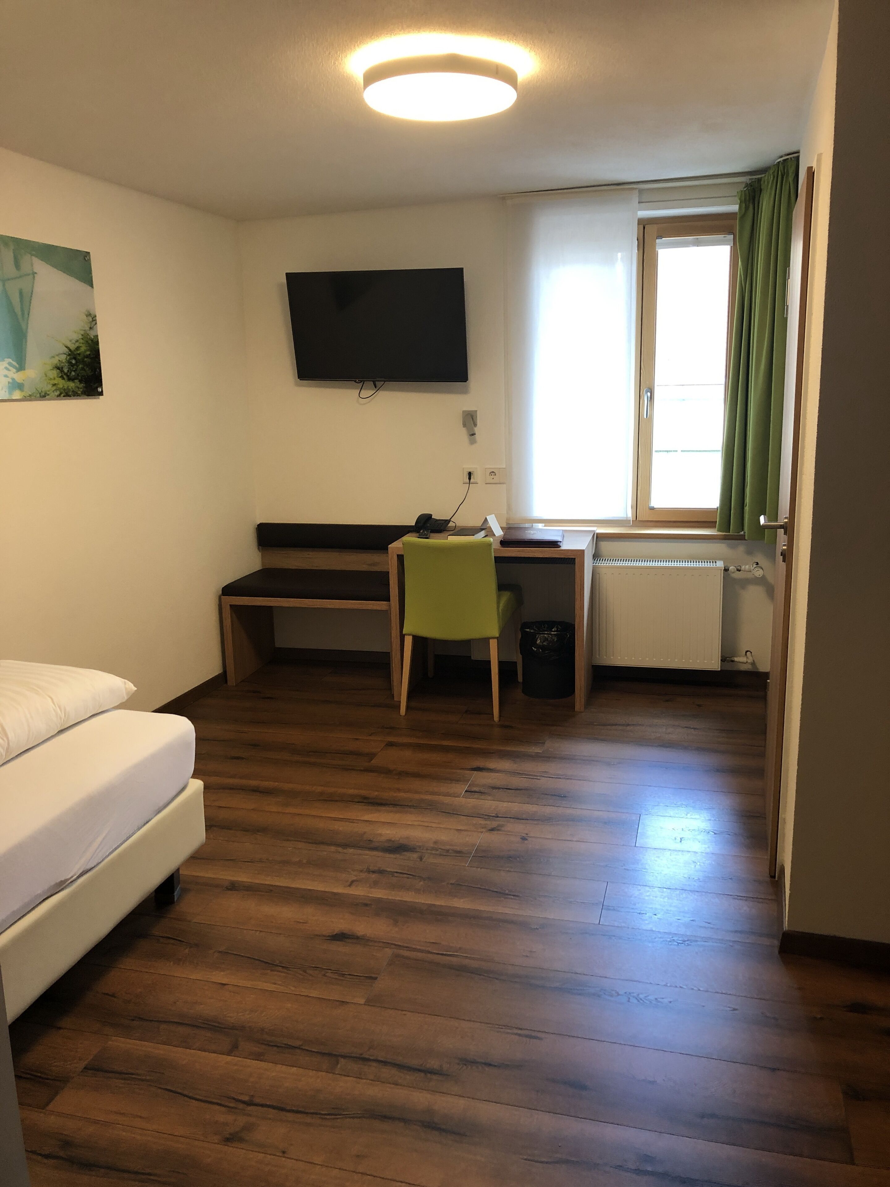 A room with a bed, a desk, a chair, a television mounted on the wall, and a window with curtains.