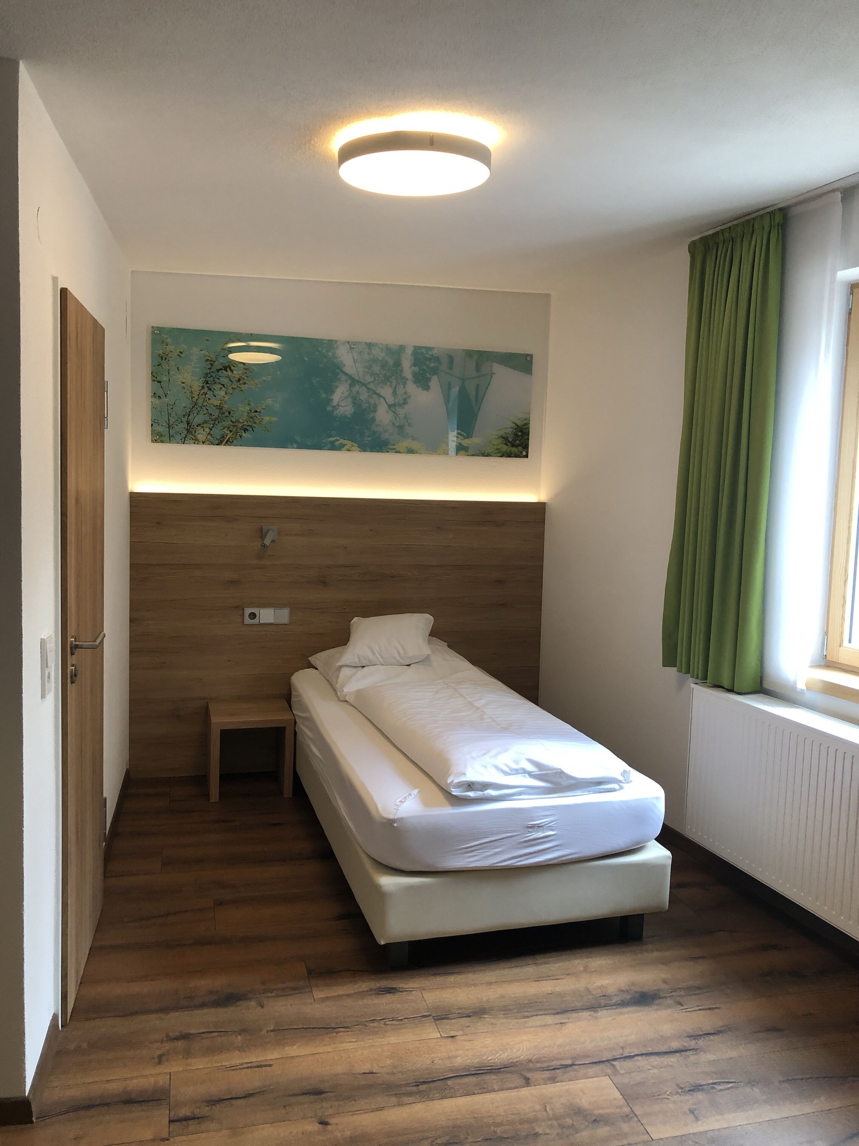 Ein kleines, ordentlich eingerichtetes Schlafzimmer mit Bett, einem kleinen Nachttisch, einer Wandleuchte, einem Gemälde und einem Fenster mit Vorhängen.