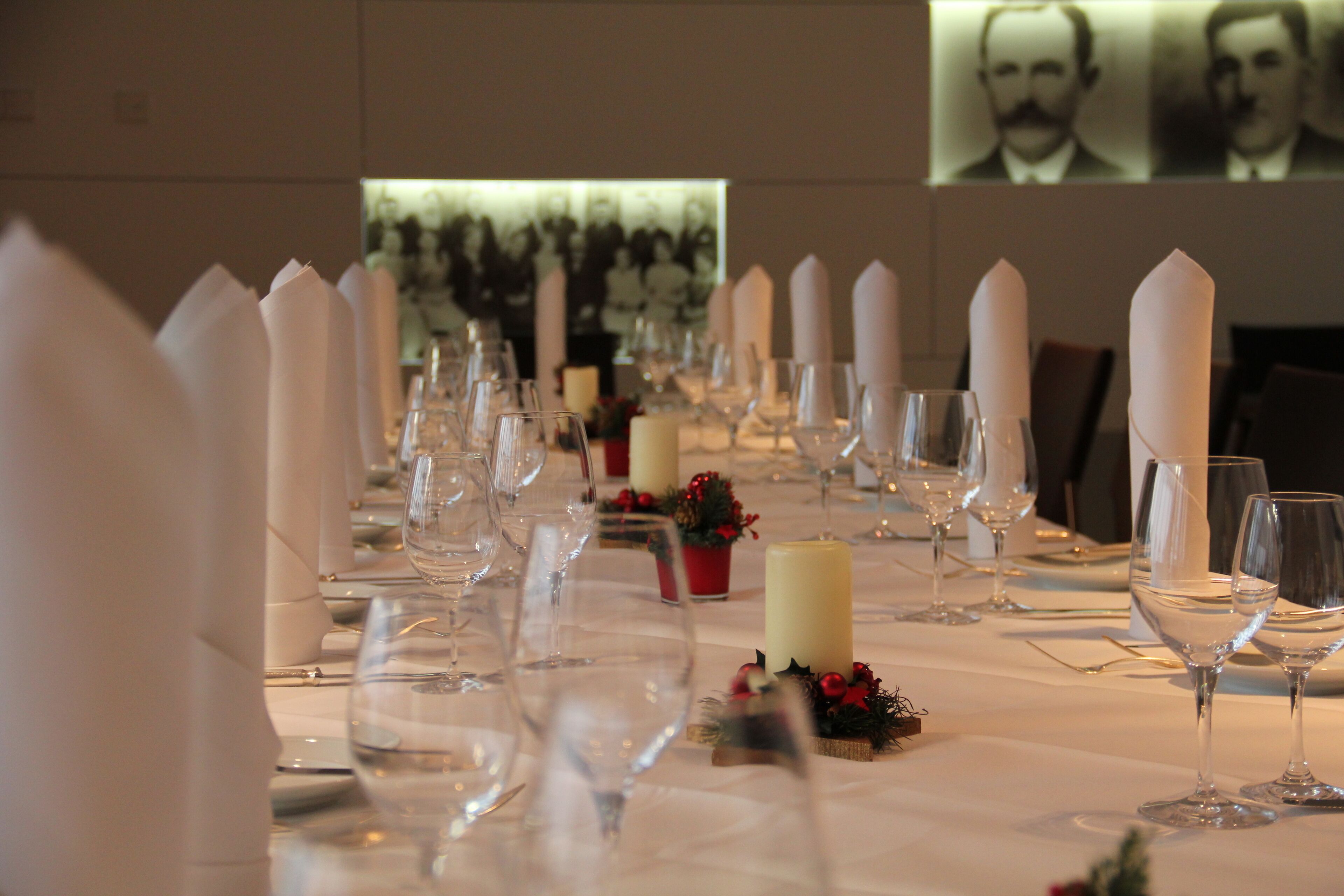 Una larga mesa de comedor dispuesta con servilletas blancas, copas de vino y velas, con una proyección de personas al fondo.
