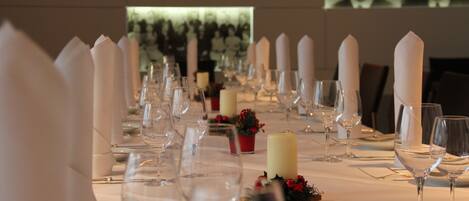 Una larga mesa de comedor dispuesta con servilletas blancas, copas de vino y velas, con una proyección de personas al fondo.