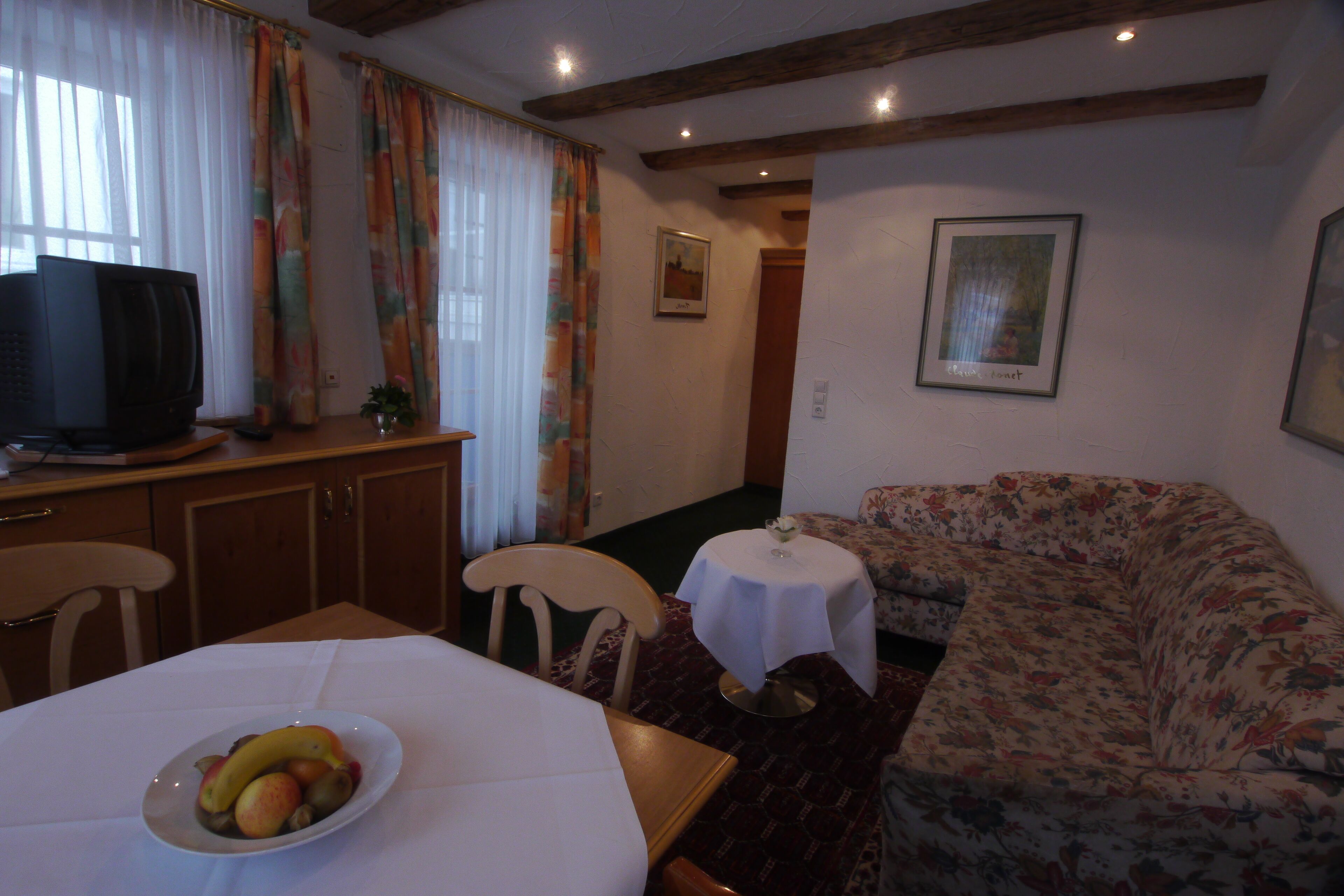 A living room with a sofa, a dining table with a fruit bowl, a television, and a small table with a flower vase.
