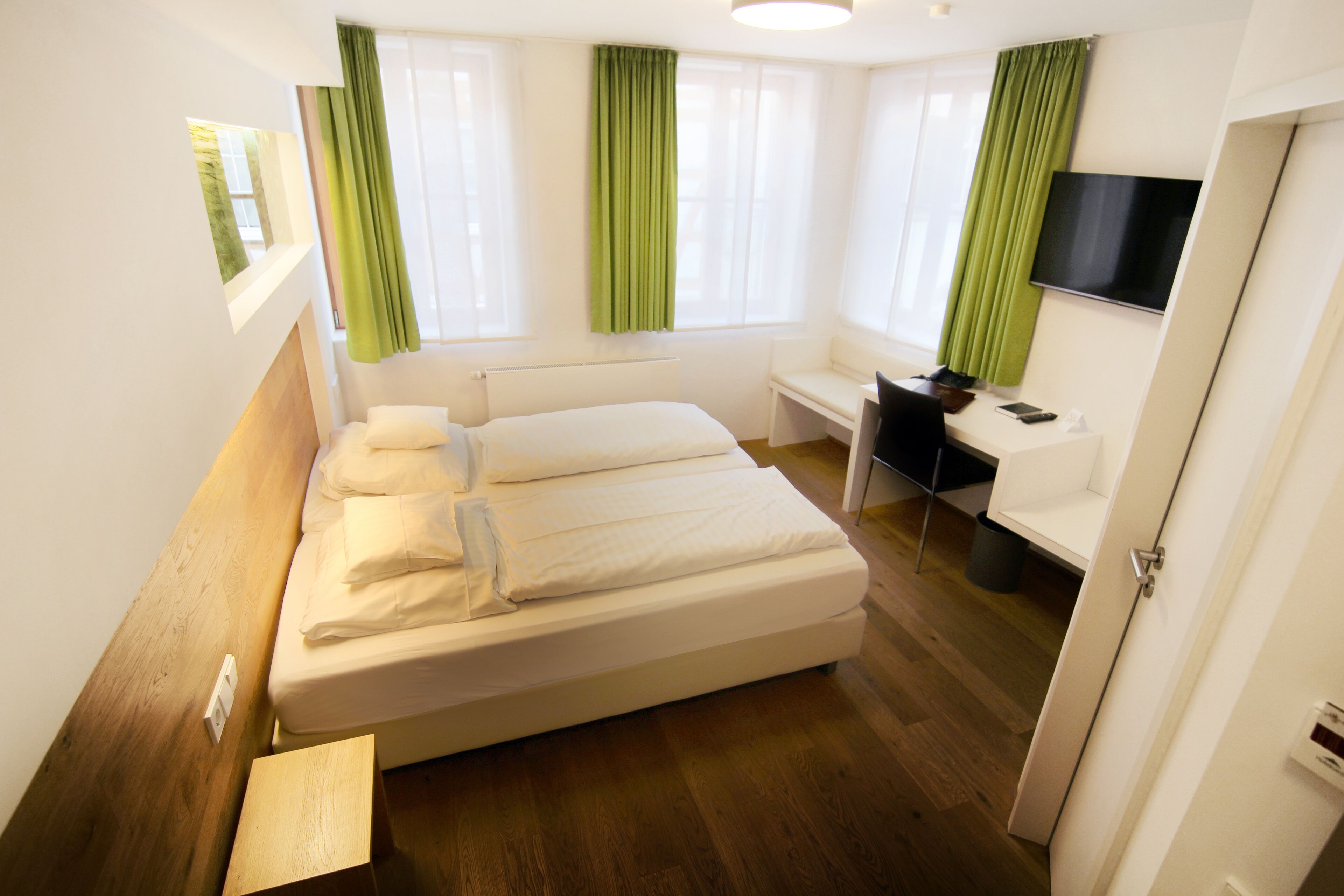 A modern hotel room with a large bed, a desk, a chair, a TV, and a window with curtains.
