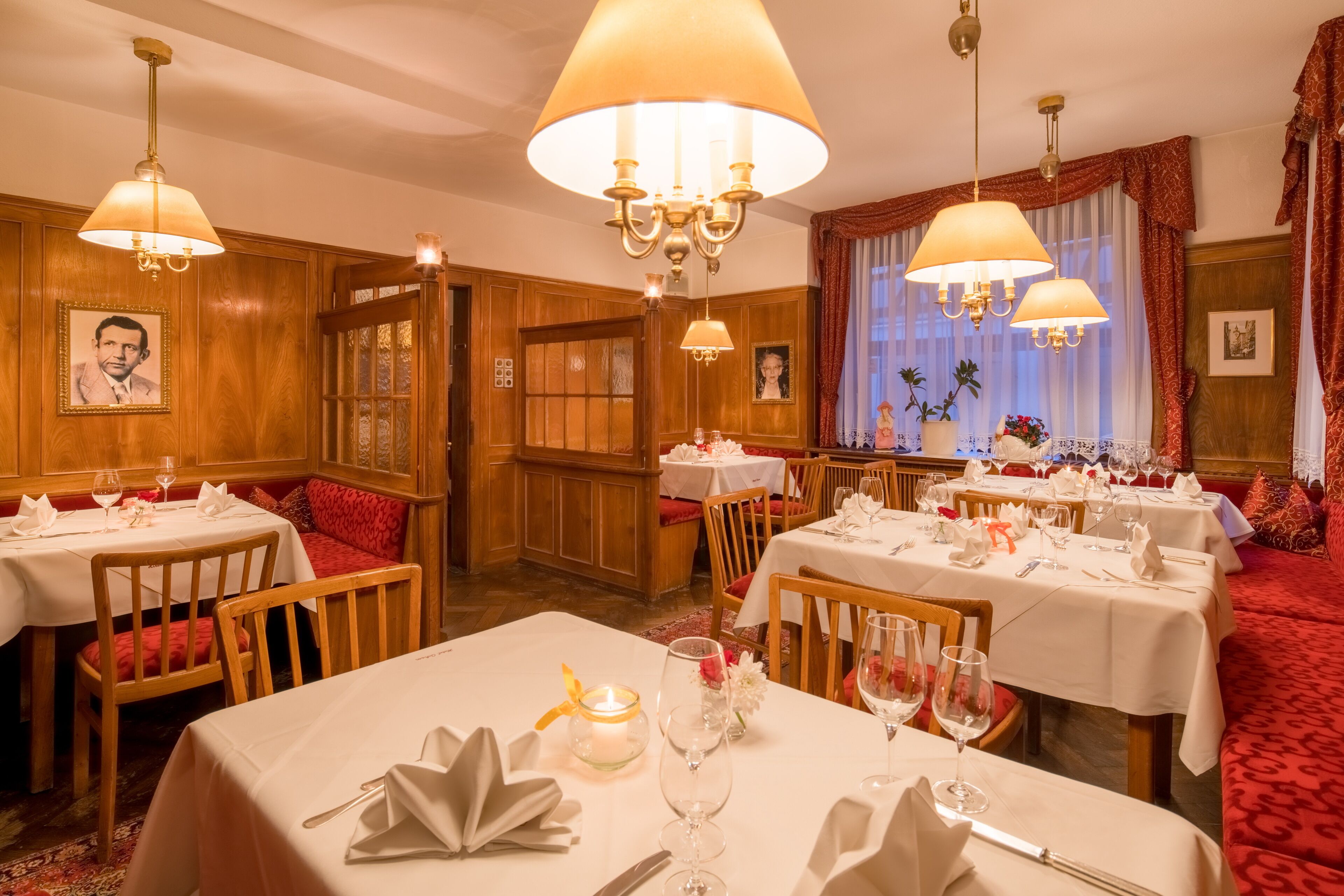 Een restaurant met houten lambrisering, kroonluchters en rode bekleding op de stoelen. Tafels zijn gedekt met witte tafellakens, wijnglazen en kaarsen.
