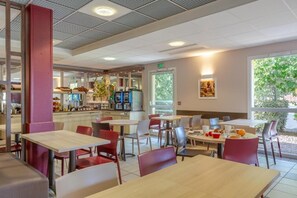 A modern dining area with tables and chairs, a kitchen in the background, and a window with a view of greenery. - Ace Hôtel Bourges (Bourges)