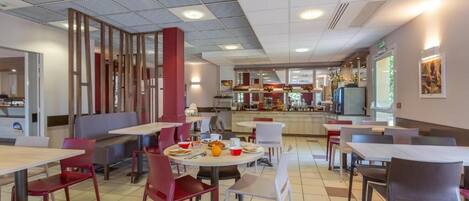 A cafeteria with tables and chairs, a counter with food items, and a kitchen area in the background.