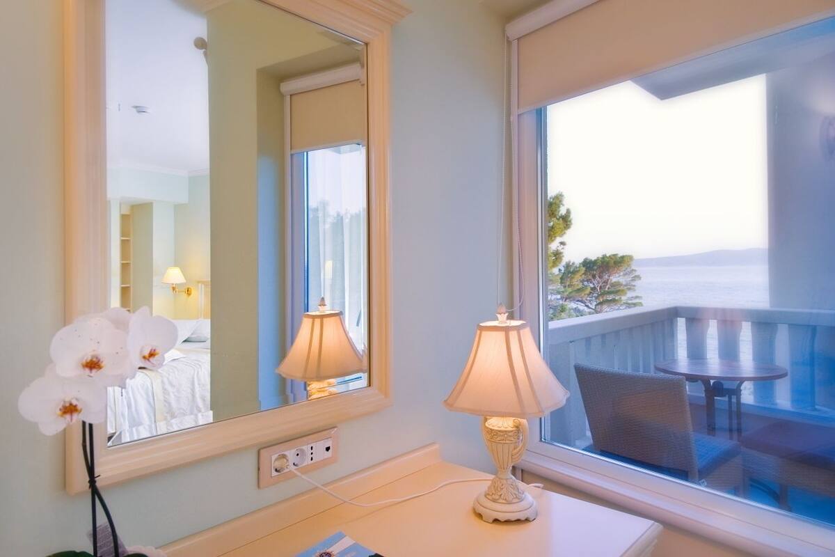 A hotel room with a large mirror, two bedside lamps, a small table with chairs, and a view of the sea through a balcony window.