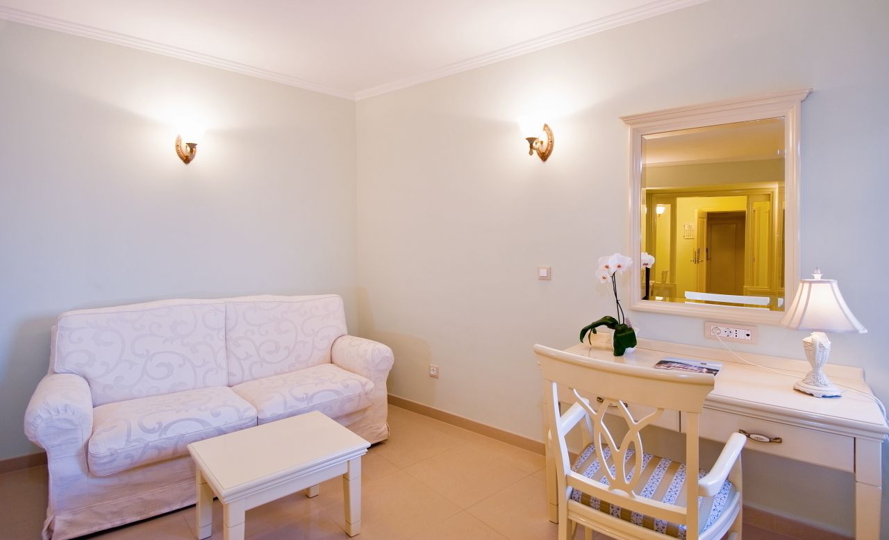 A small, well-lit room with a white sofa, a small table, a chair, a desk with a lamp, and a mirror.