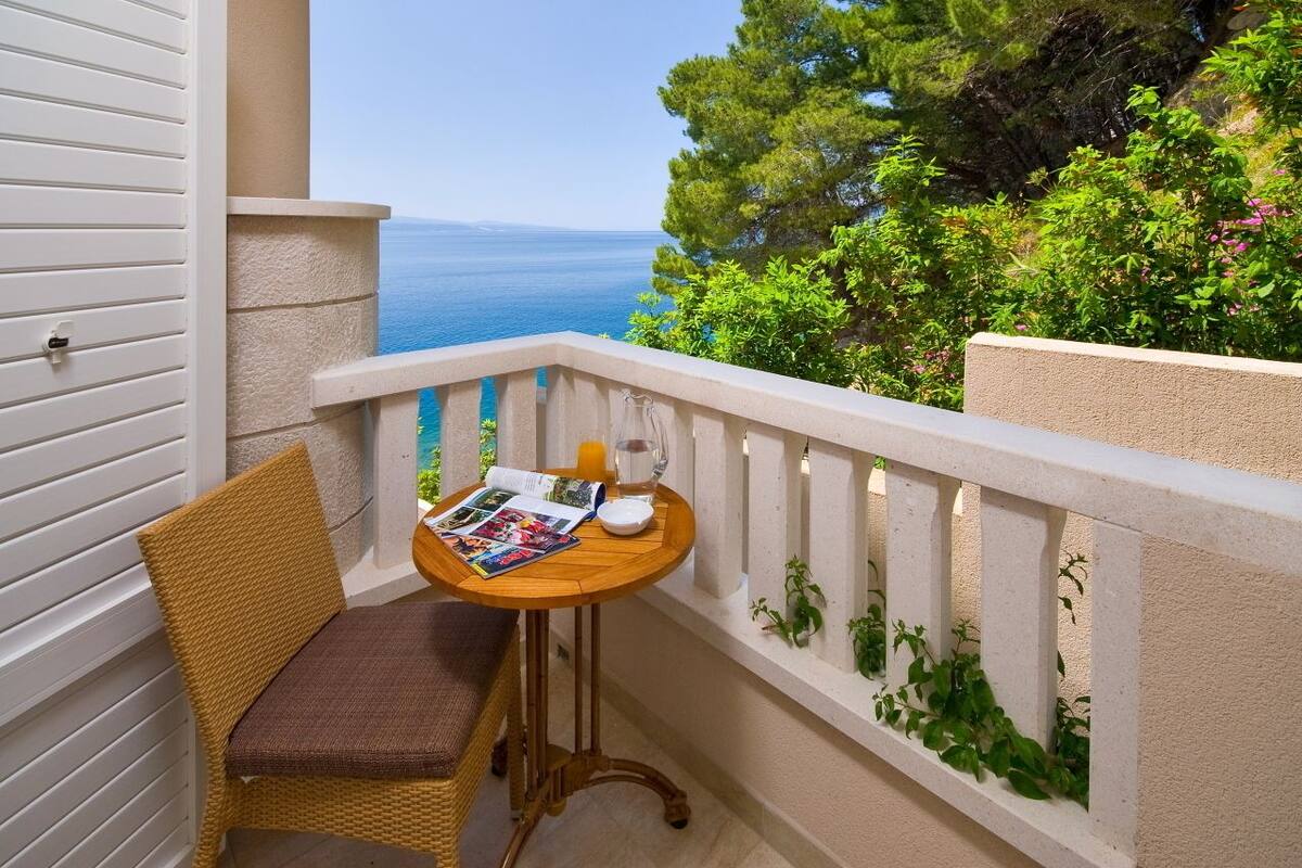 A balcony with a wicker chair, a small round table with a magazine, a glass of orange juice, and a view of the sea.