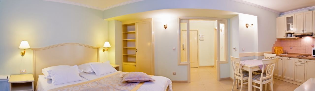 A bedroom with a bed, bedside tables, a desk, and a chair. There is a kitchen area with cabinets and a countertop.