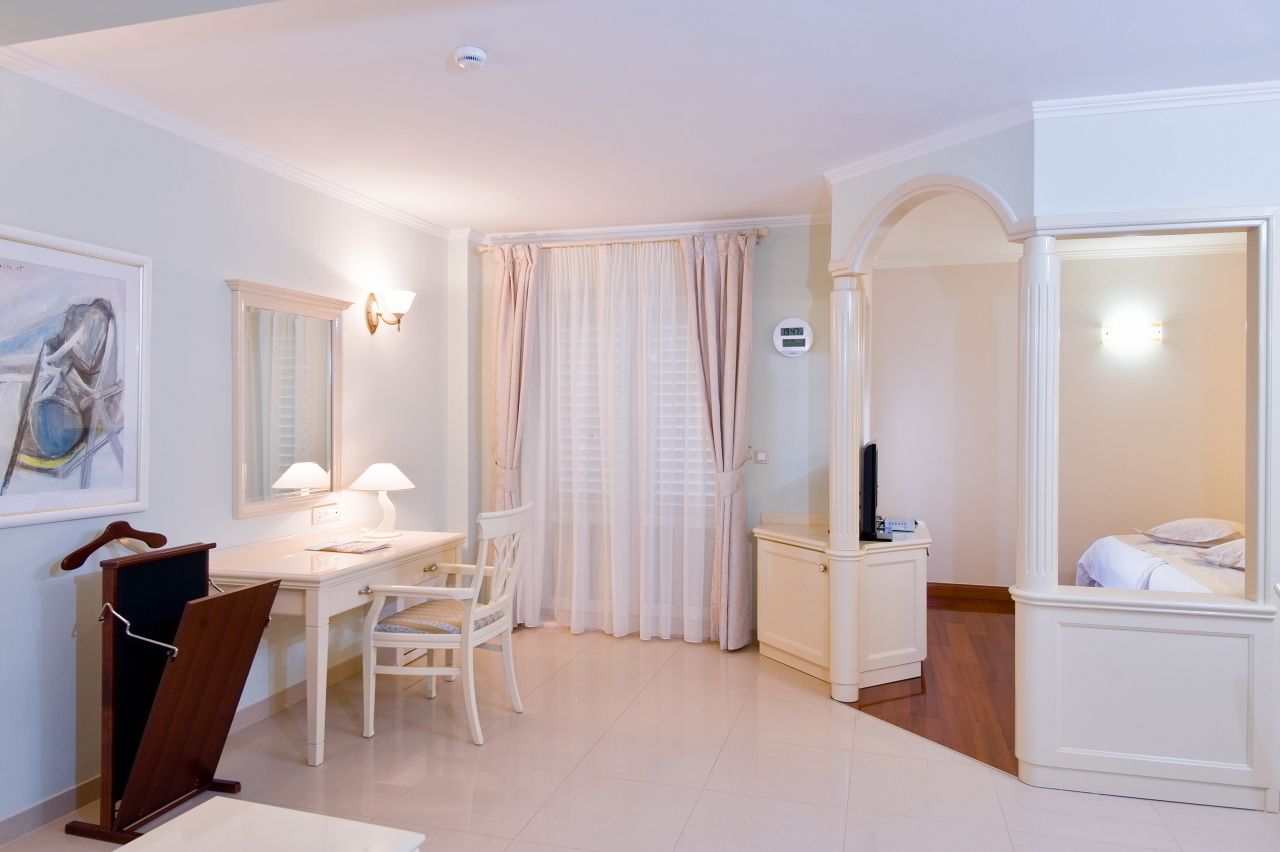 A hotel room with a desk, chair, and a bed. There is a window with curtains, a wall-mounted TV, and a painting on the wall.