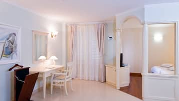 A hotel room with a desk, chair, and a bed. There is a window with curtains, a wall-mounted TV, and a painting on the wall.