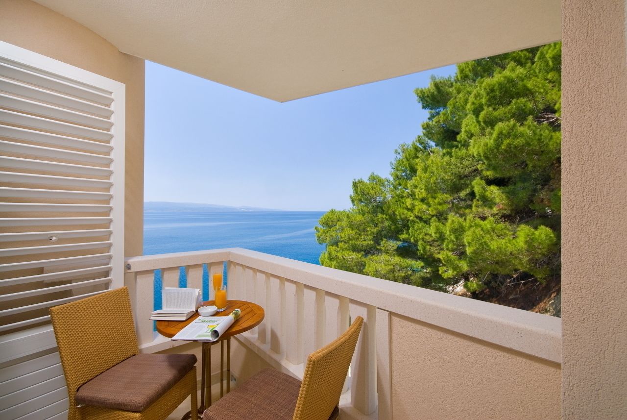 A balcony with two wicker chairs and a small table, overlooking a coastal view with trees and the sea.