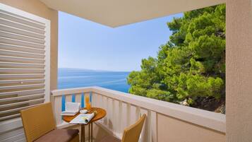 A balcony with two wicker chairs and a small table, overlooking a coastal view with trees and the sea.