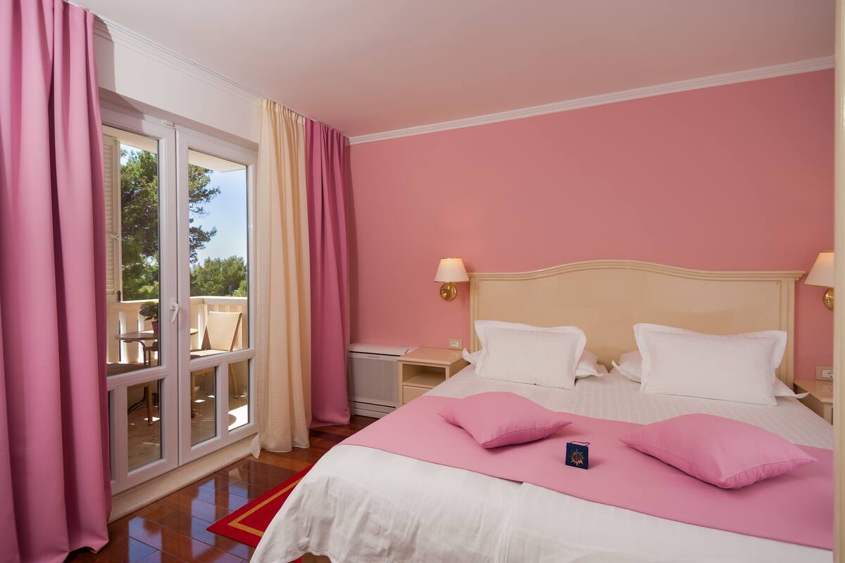 A bedroom with a large bed, a nightstand, a lamp, and a view through a glass door to an outdoor area with a table and chairs.