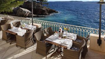 A seaside restaurant terrace with wicker furniture and white tablecloths, overlooking the ocean.