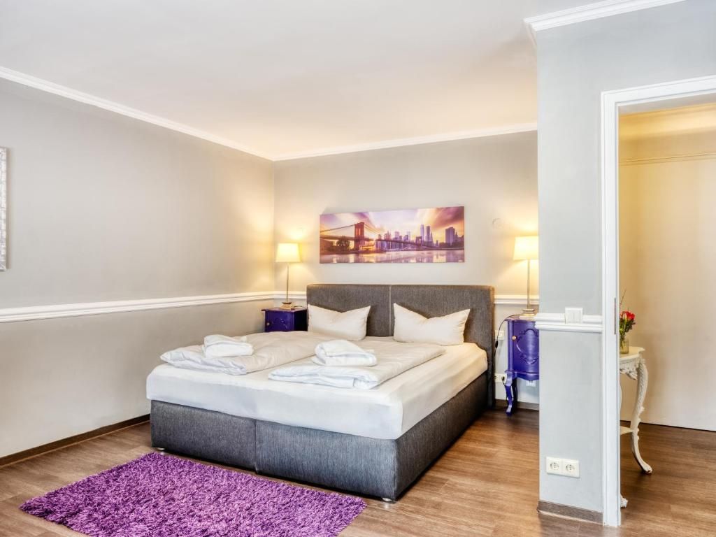 Habitación de hotel con una cama, una alfombra púrpura, un cuadro en la pared y una mesita de noche con una lámpara.
