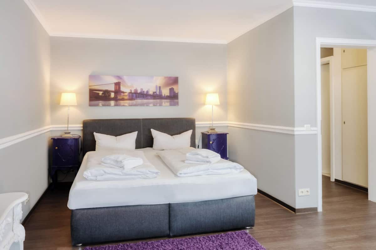 Ein Hotelzimmer mit einem großen Bett, zwei Nachttischen, einem Stadtbild an der Wand und einem violetten Teppich.