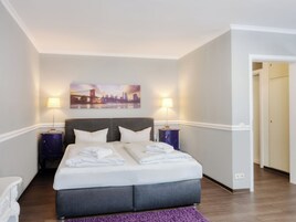 Una habitación de hotel con una cama grande, dos mesitas de noche, un cuadro de un paisaje urbano en la pared y una alfombra morada.