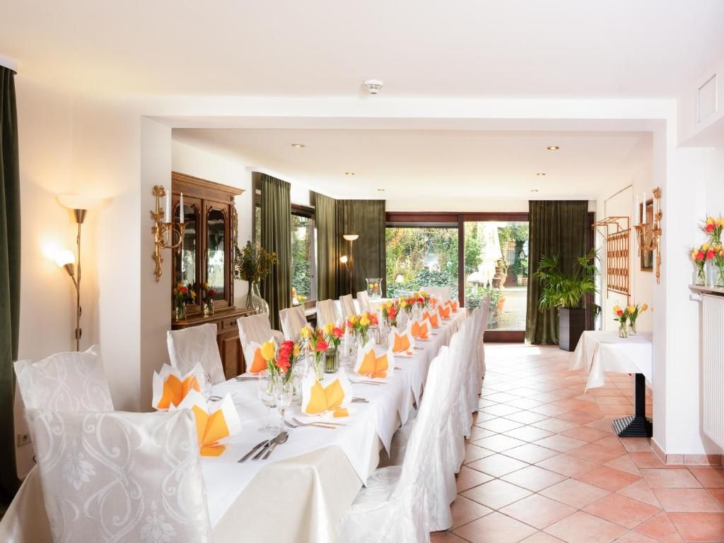 Une longue table à manger, dressée pour un repas avec des nappes blanches et des serviettes orange, entourée de chaises recouvertes de housses blanches, dans une pièce au sol carrelé et offrant une vue sur la verdure à l’extérieur.