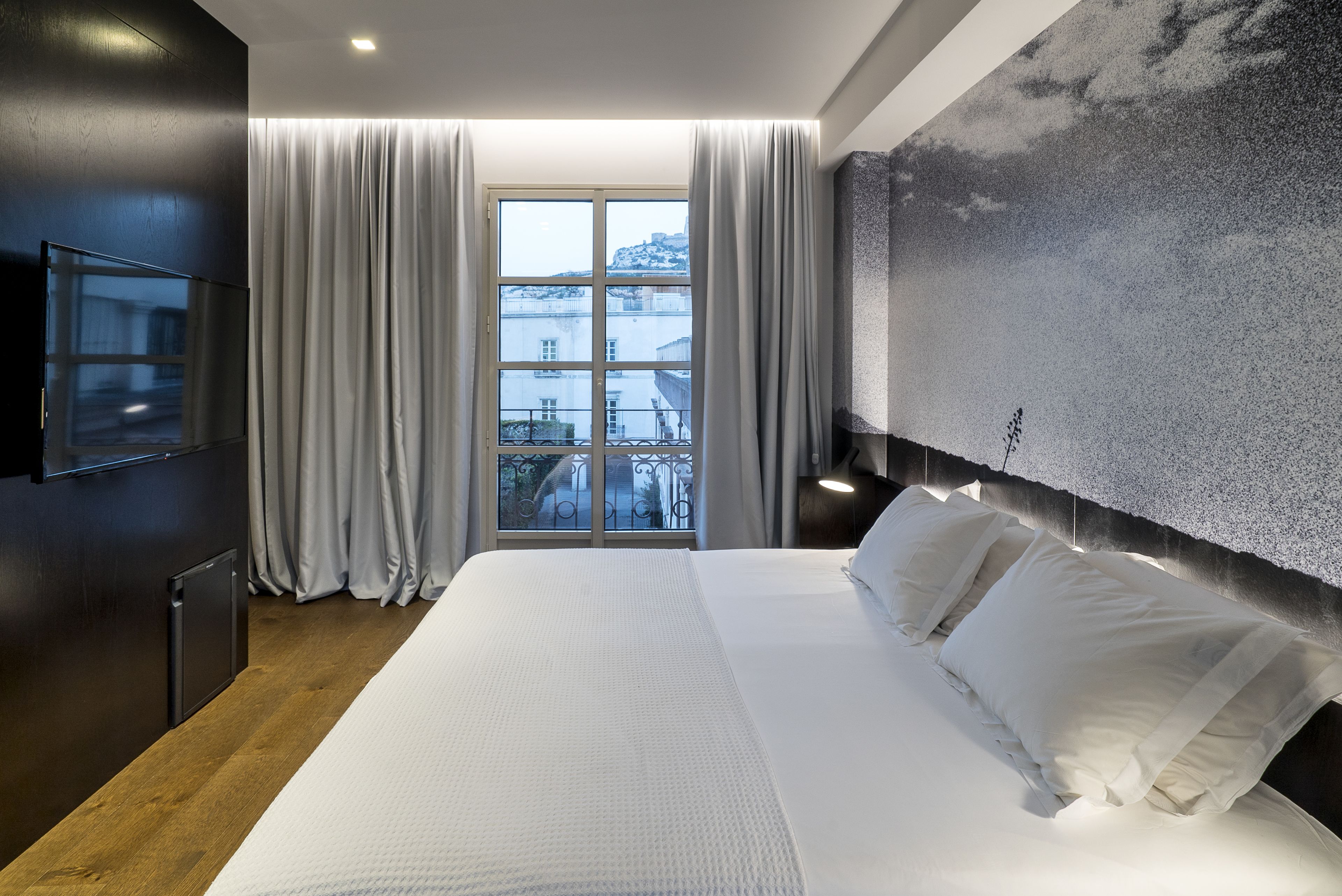A modern hotel room with a large bed, a flat-screen TV, a view of the city through the window, and a textured wall.