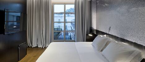 A modern hotel room with a large bed, a flat-screen TV, a view of the city through the window, and a textured wall.