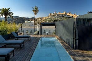 Una piscina en la azotea con sillas de descanso, una terraza de madera y vistas a una fortaleza histórica en la colina.