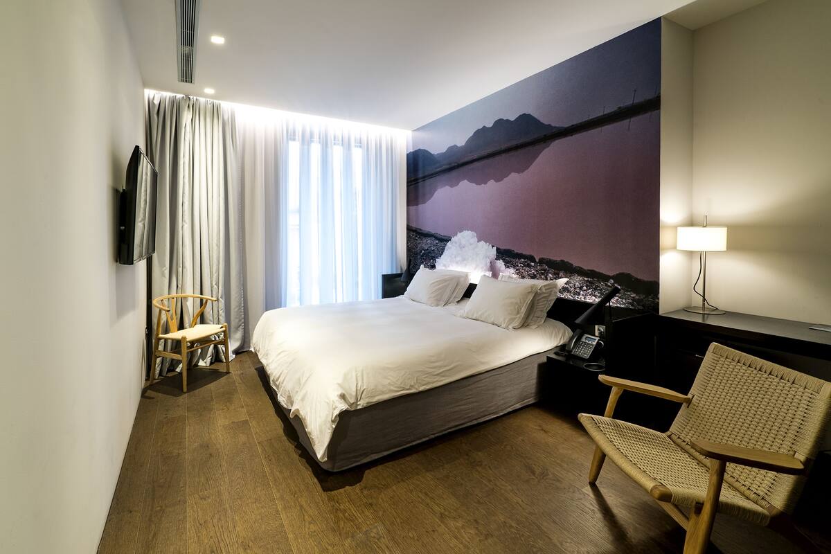 A modern hotel room with a large bed, a desk with a lamp, a chair, and a wall-mounted TV.