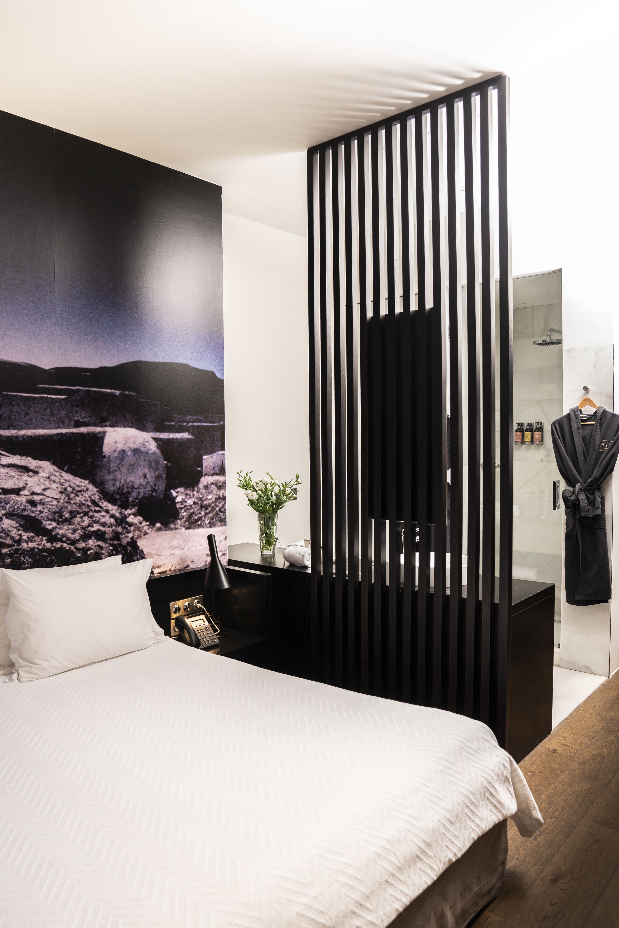 A modern hotel room with a large bed, a black partition, a mounted TV, and a bathroom with a glass shower.