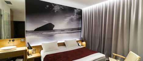 A modern hotel room with a large bed, a wooden nightstand, a round mirror, a bedside table with a lamp, and a large wall mural of a beach scene.