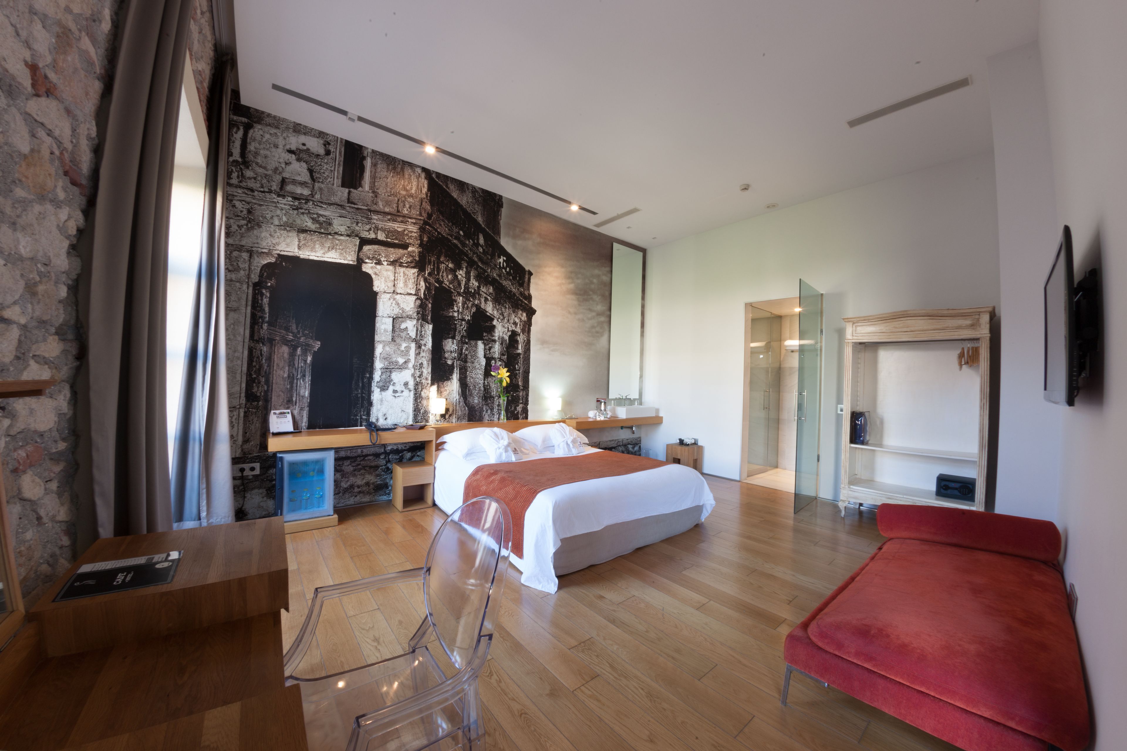 A modern bedroom with a stone wall, wooden flooring, a bed, a red chair, and a TV.