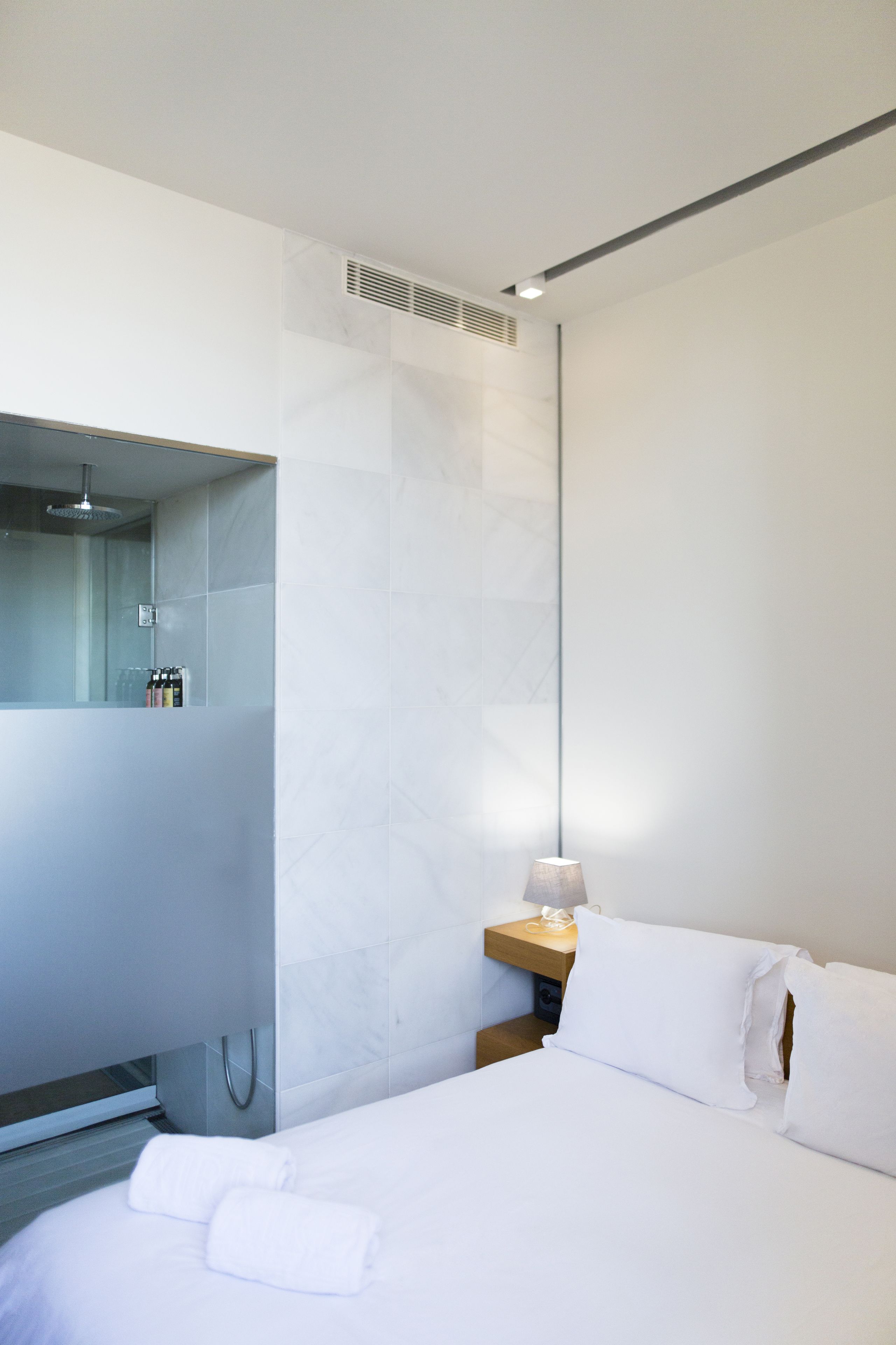 A modern hotel room with a large bed, a bedside table with a lamp, a wooden nightstand, and a built-in shower area with a glass partition.