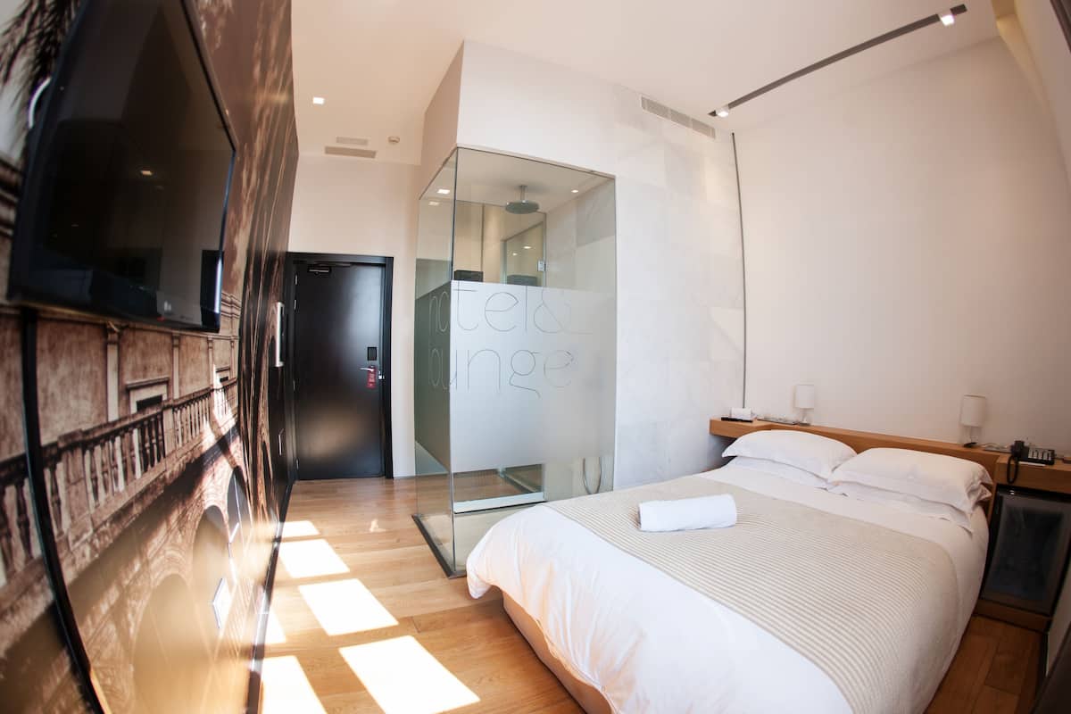 A modern hotel room with a bed, bedside lamps, a TV, and a glass partition with 'hotel lounge' written on it.