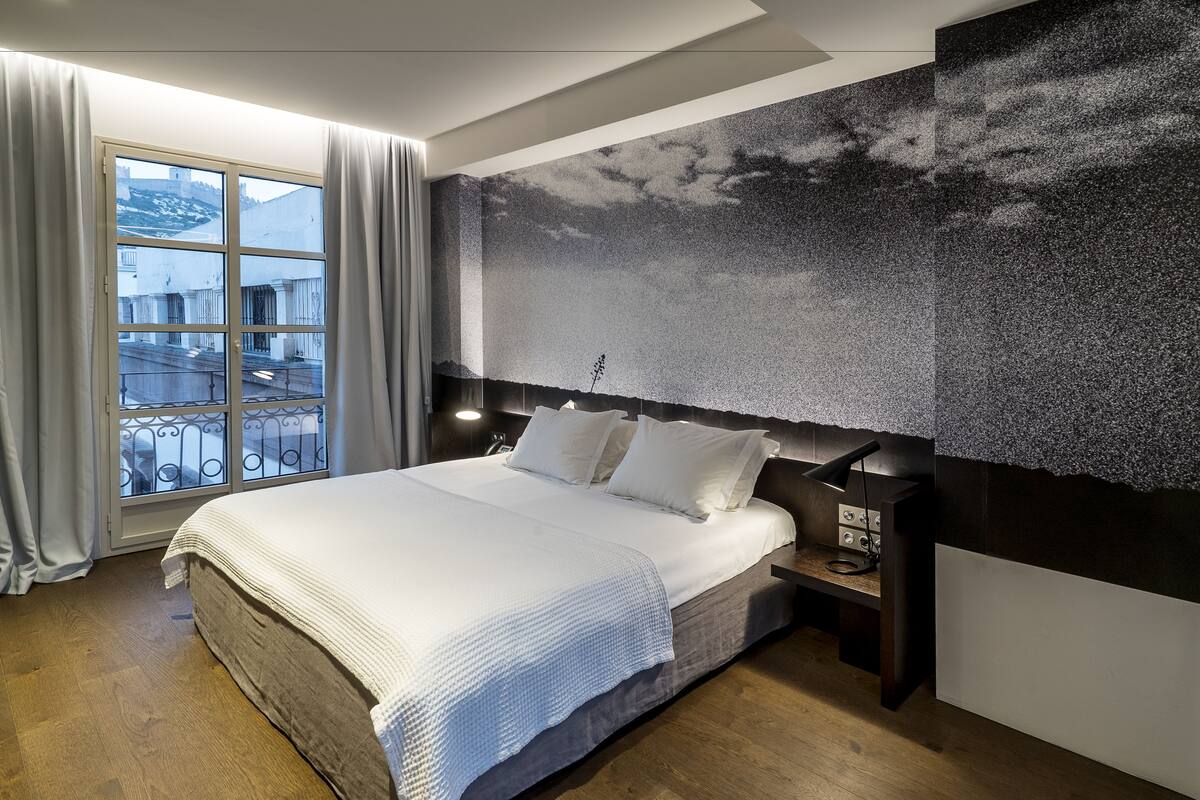 A modern bedroom with a large bed, a view of buildings through a window, and a textured wall.