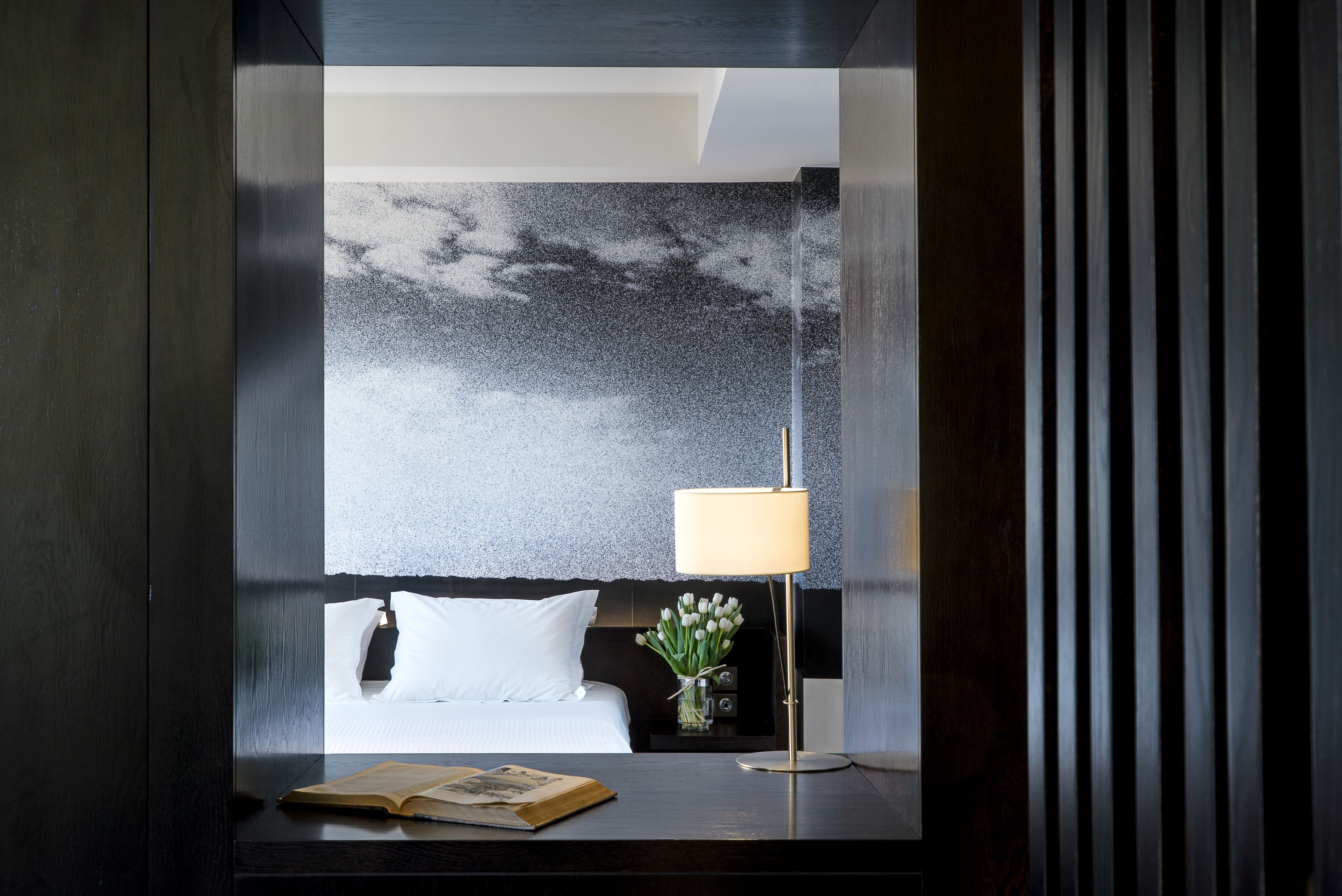 Una habitación de hotel moderna con un ventanal grande, una cama con sábanas blancas, una mesita de noche con una lámpara y un florero, y un libro sobre la mesa.