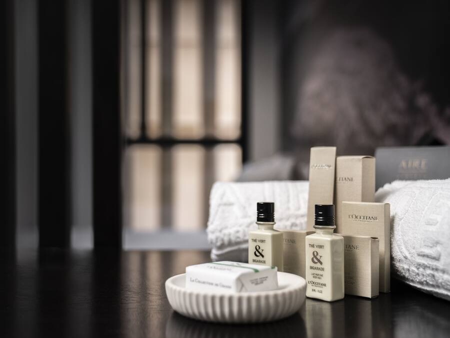A neatly arranged set of toiletries on a wooden surface, including a bottle, bar of soap, and boxes, with a blurred background suggesting a room setting.