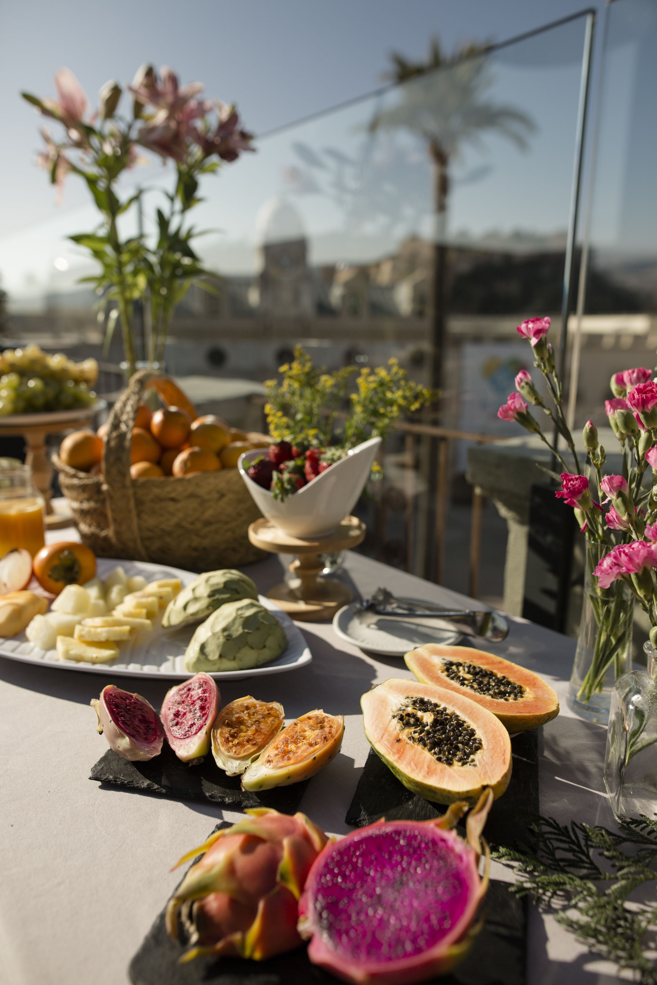 Un tavolo con diversi tipi di frutta e verdura, tra cui papaya e pitaya, su un balcone con vista su edifici e palme.