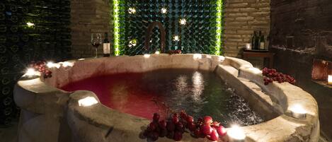 Una bañera de piedra con vino tinto y uvas, rodeada de un muro de ladrillo e iluminada por velas.