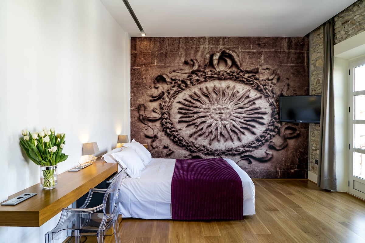 A modern hotel room with a large wall mural, a wooden desk, a bed with a purple throw, and a TV on the wall.