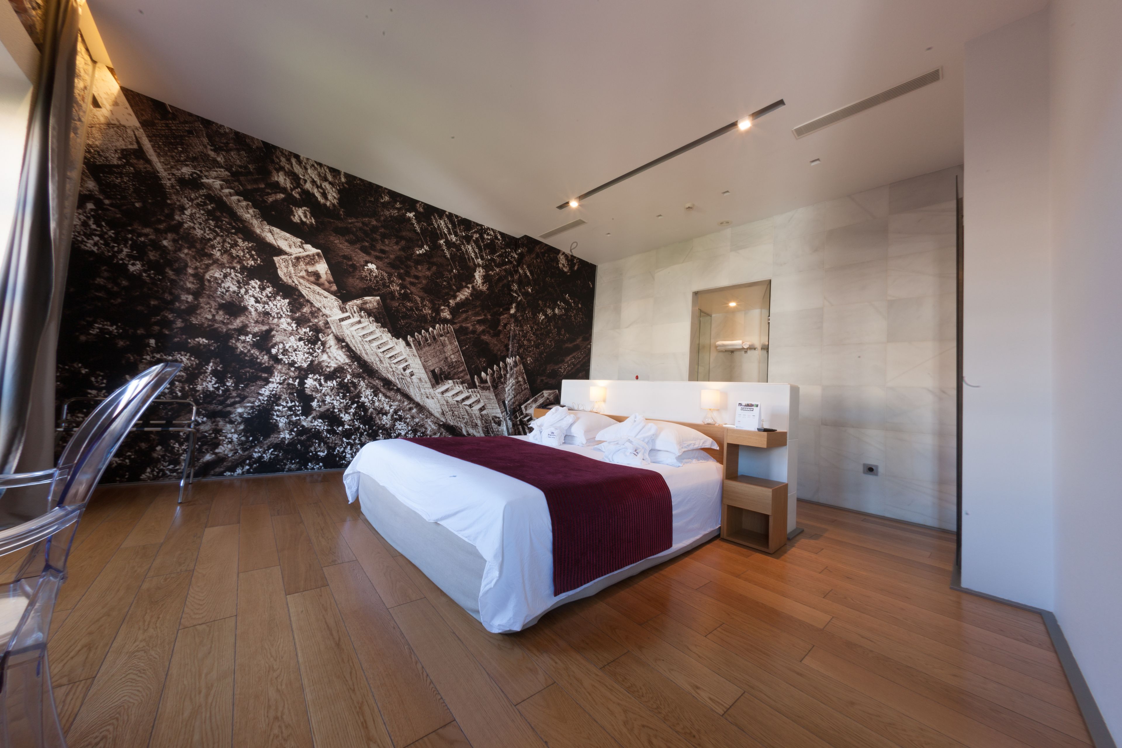 Un dormitorio moderno con una cama grande, un mural de mármol, piso de madera y una silla de vidrio.