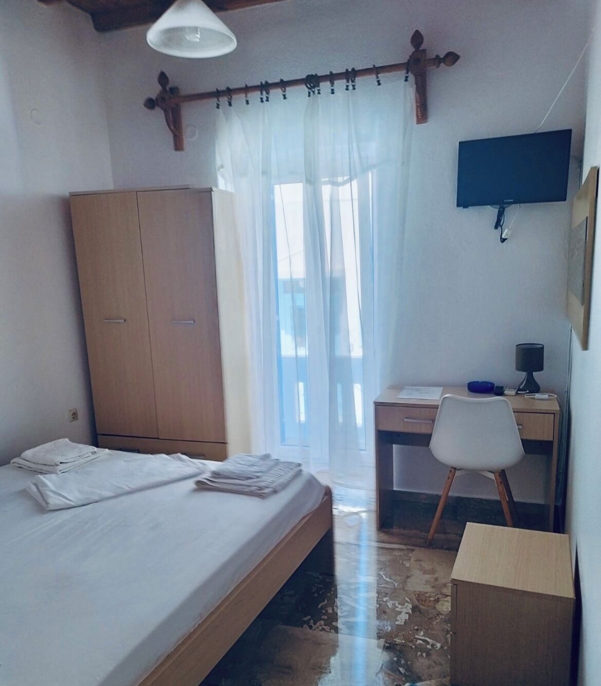 A bedroom with a bed, a wooden wardrobe, a desk with a chair, a television, and a window with sheer curtains.