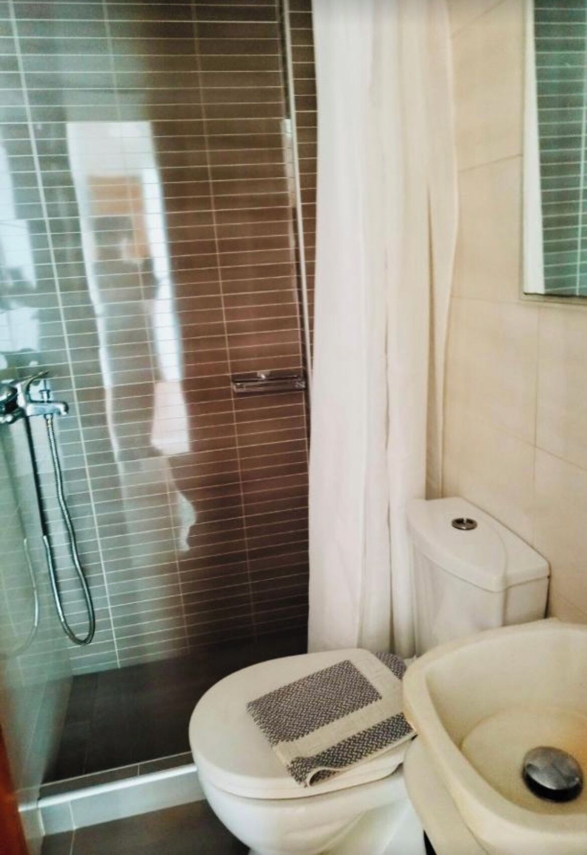 Une salle de bain avec une douche à parois de verre, des toilettes surmontées d’un tapis à motifs et un lavabo avec un drain.