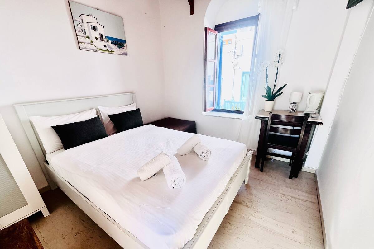 A neatly made bed with white linens and pillows, a bedside table with a plant, a window with curtains, and a painting on the wall.