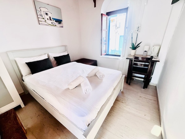 A neatly made bed with white linens and pillows, a bedside table with a plant, a window with curtains, and a painting on the wall.
