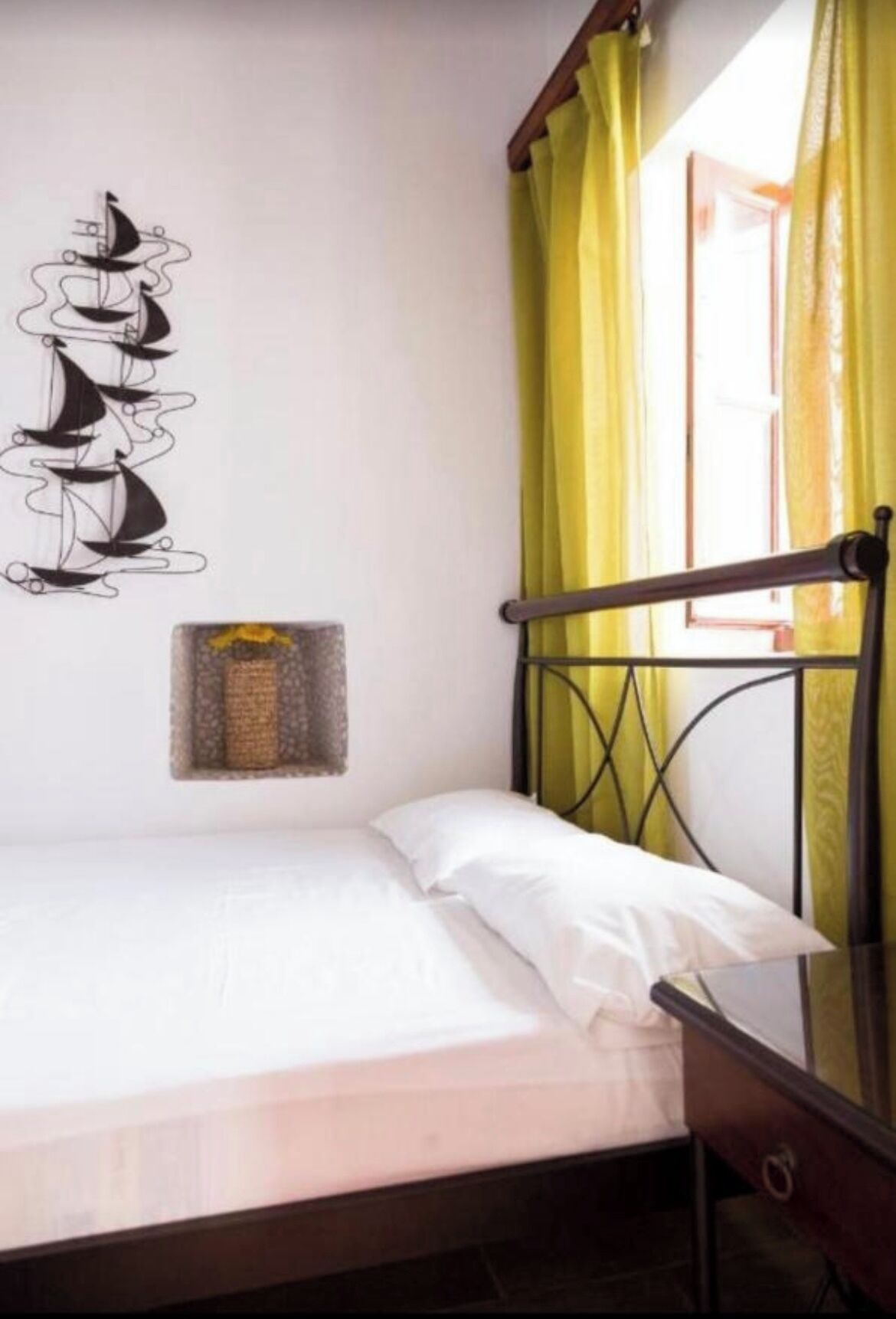 A single bed with a metal frame, a window with yellow curtains, and a wall decoration of sailboats.