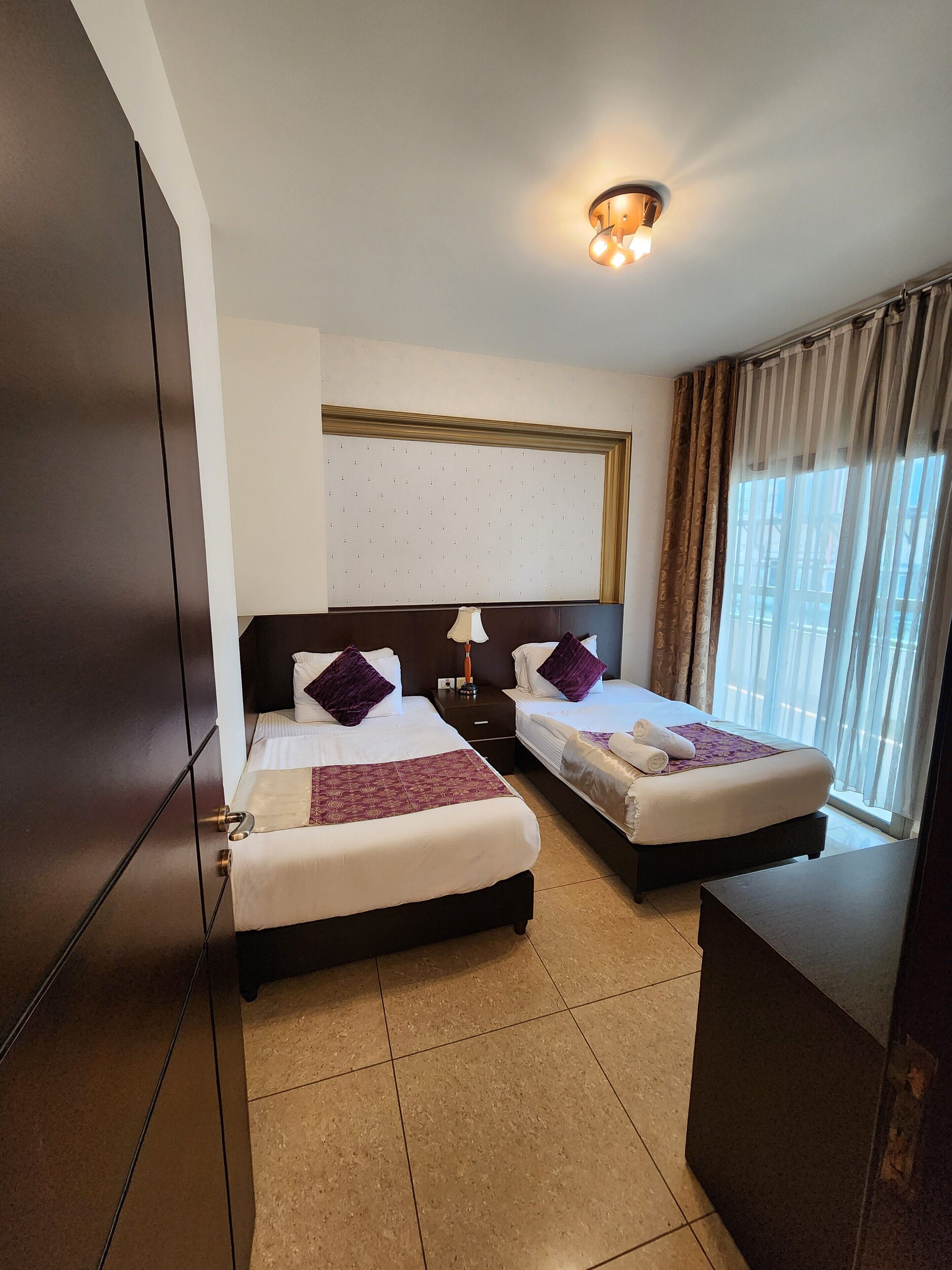 A hotel room with two beds, a large mirror, a ceiling light, and a window with curtains.