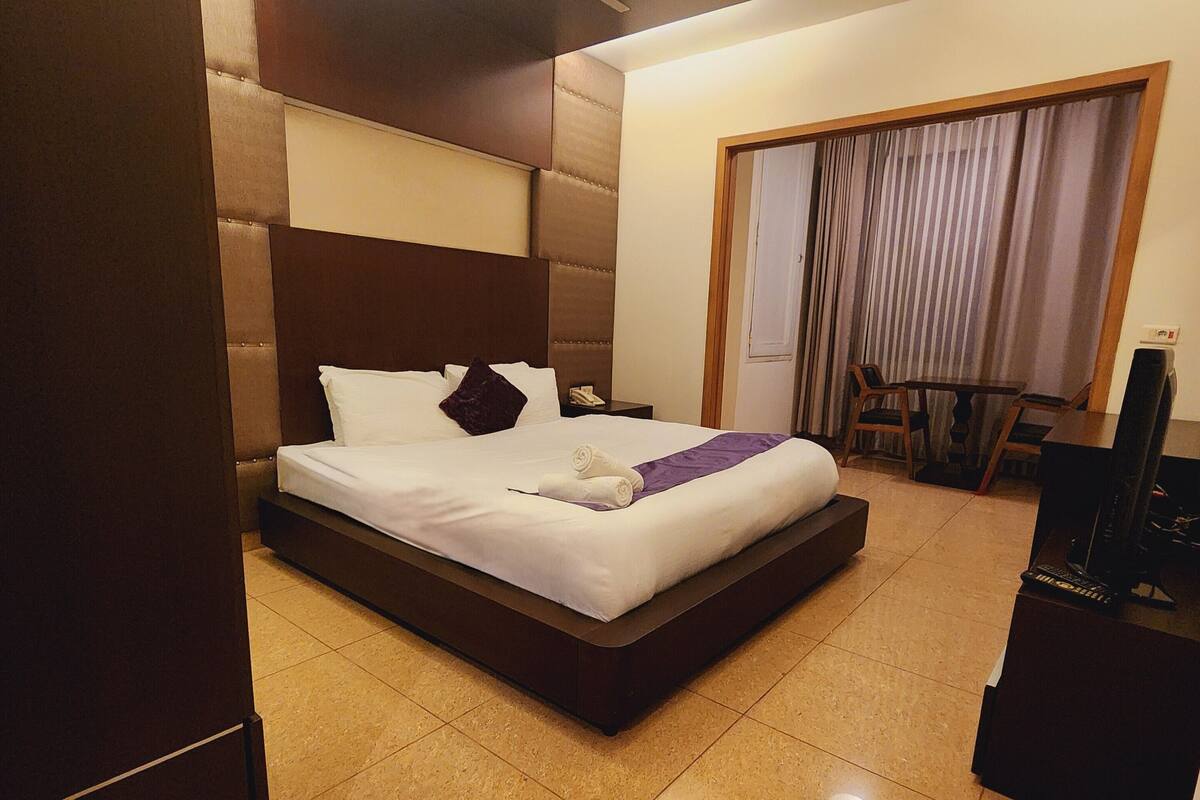 Habitación de hotel con una cama grande, una mesita con sillas, televisión y una puerta que da a un balcón.