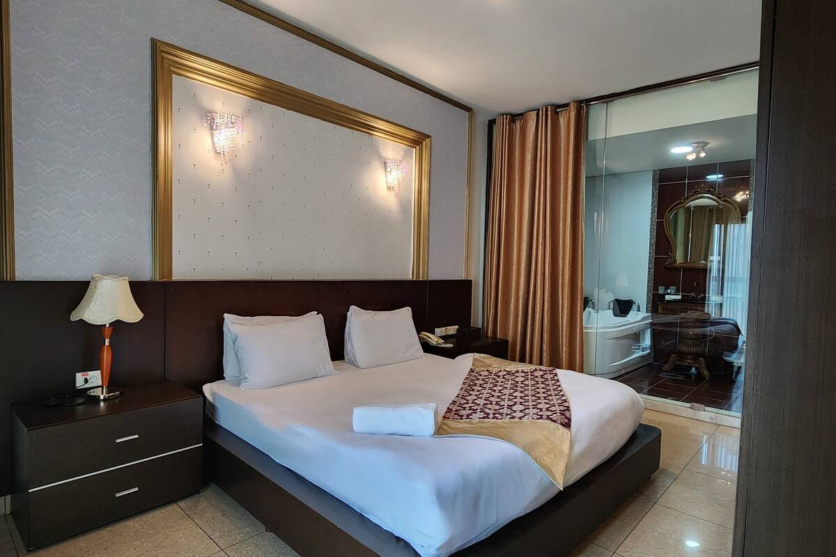 Habitación de hotel con una cama grande, una mesita de noche con lámpara, un aplique de pared y un espejo.