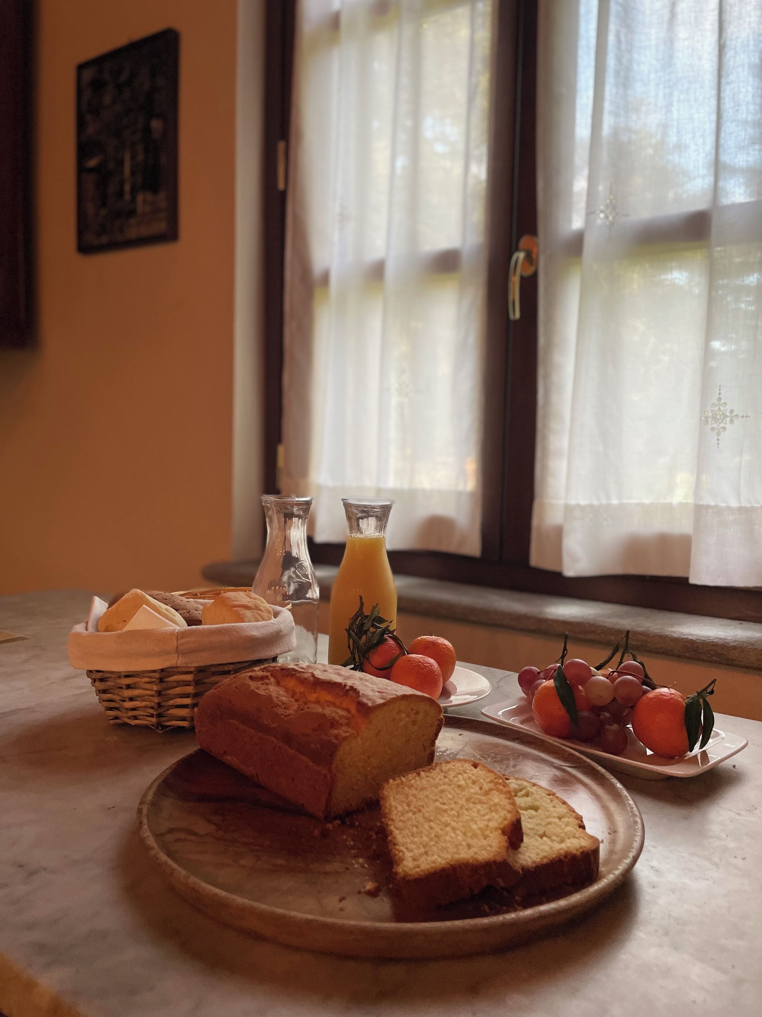Un desayuno con pan, una botella de jugo de naranja y fruta fresca sobre una mesa cerca de una ventana con cortinas.